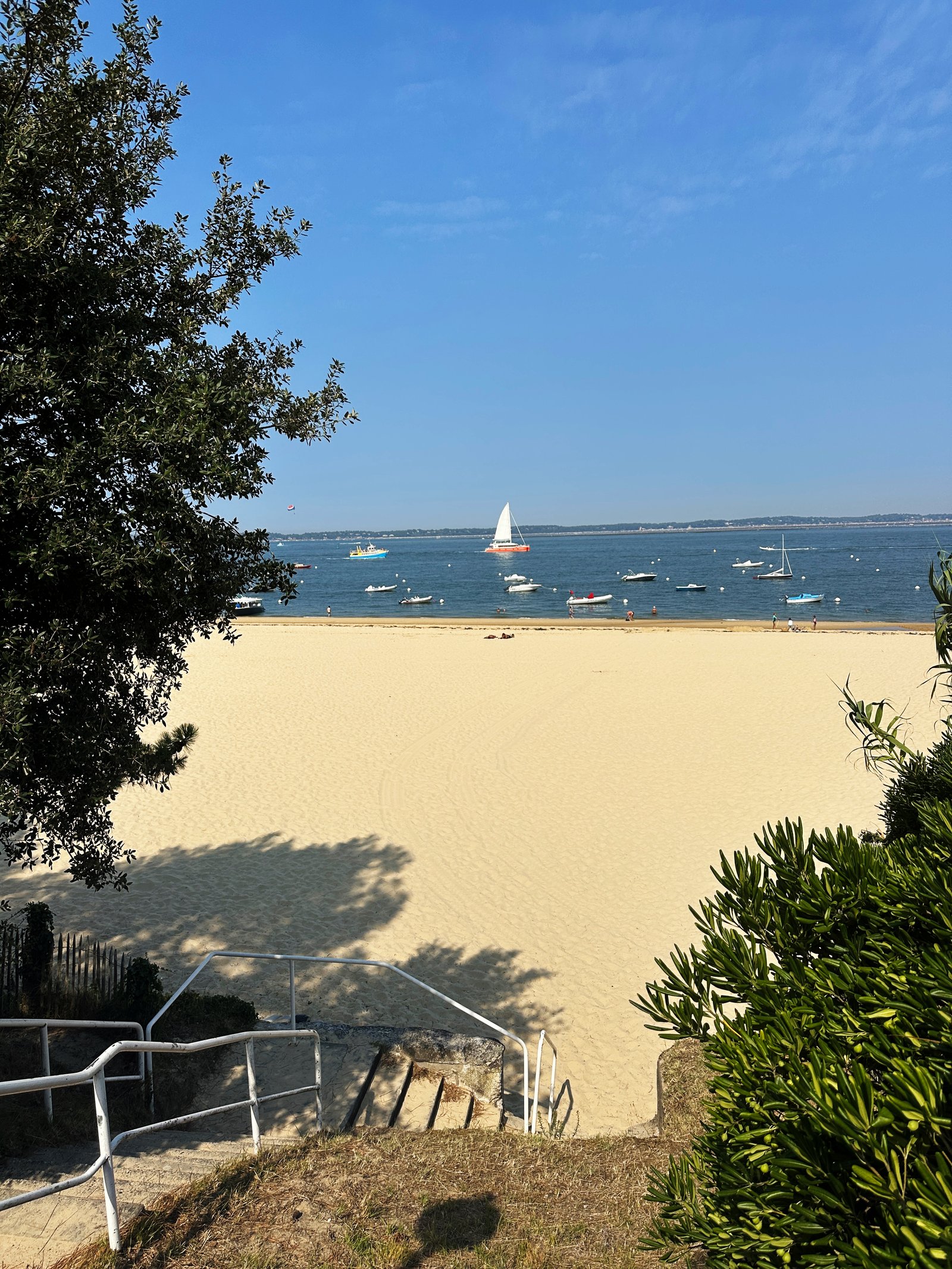 [Image de la descente d'escaliers en bois vers la plage de sable fin d'Arcachon, face aux bateaux sur l'eau bleue]